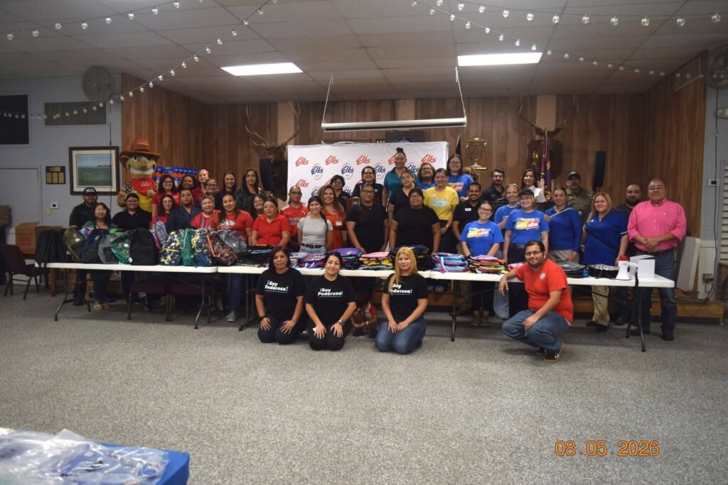 Around forty Operation Christmas RGV volunteers standing behind long tables filled with backpacks and school supplies prepared for local families